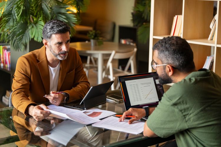 Two men discussing documents and data on laptops in an office.