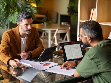Two men discussing data and charts at a glass table in an office.