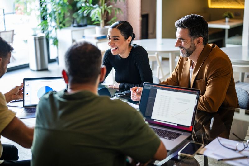 Iranian businesswoman and male co-workers sitting at table with laptops, brainstorming ideas, enthusiasm, energy, positivity