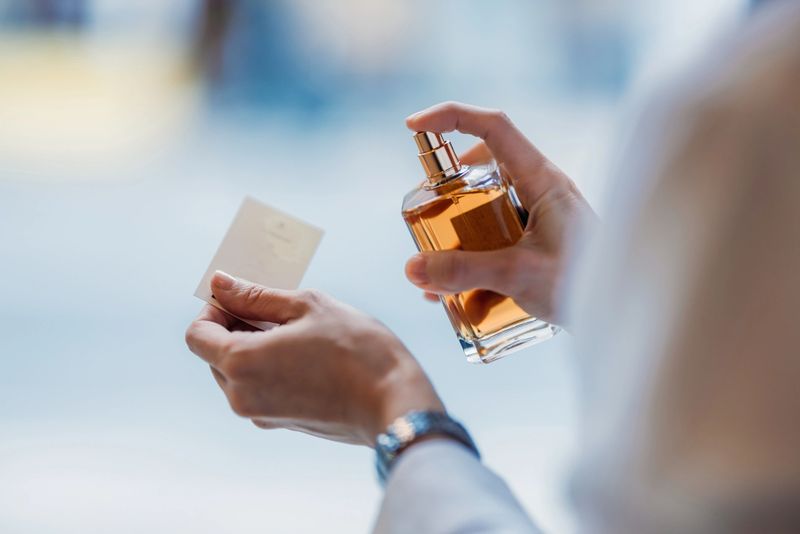 A hand sprays a perfume bottle onto a testing card, trying the fragrance in closeup. The scene shows a focused moment of scent testing and evaluation in a professional or personal setting.