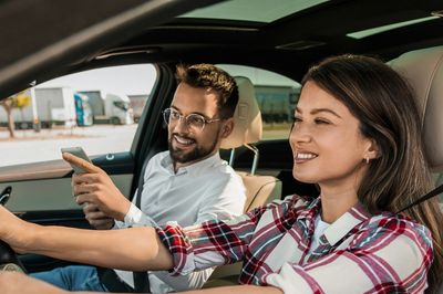 Smiling woman driving car with man showing something on phone.