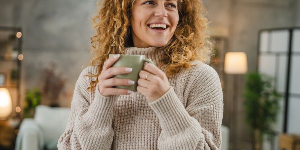 Smiling woman with curly hair holding a mug in a cozy sweater indoors.