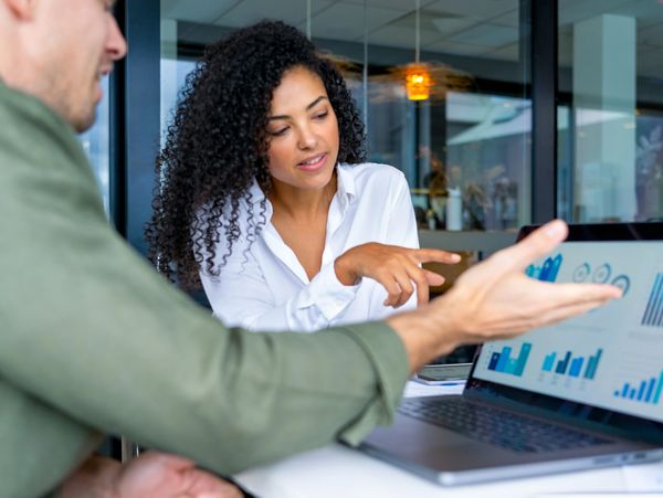 Two colleagues discussing data charts displayed on a laptop screen in an office.