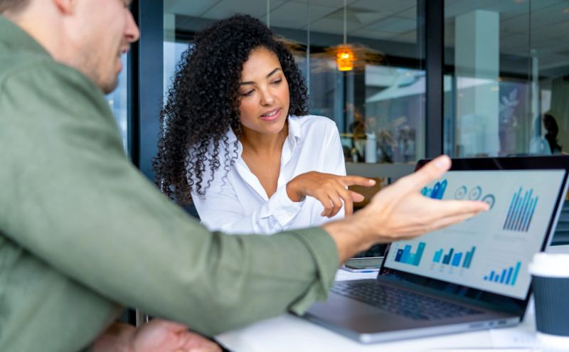 Business man and business woman in a meeting at the office. There is a laptop on the table  showing finance charts and graphs. They are looking and pointing at the computer