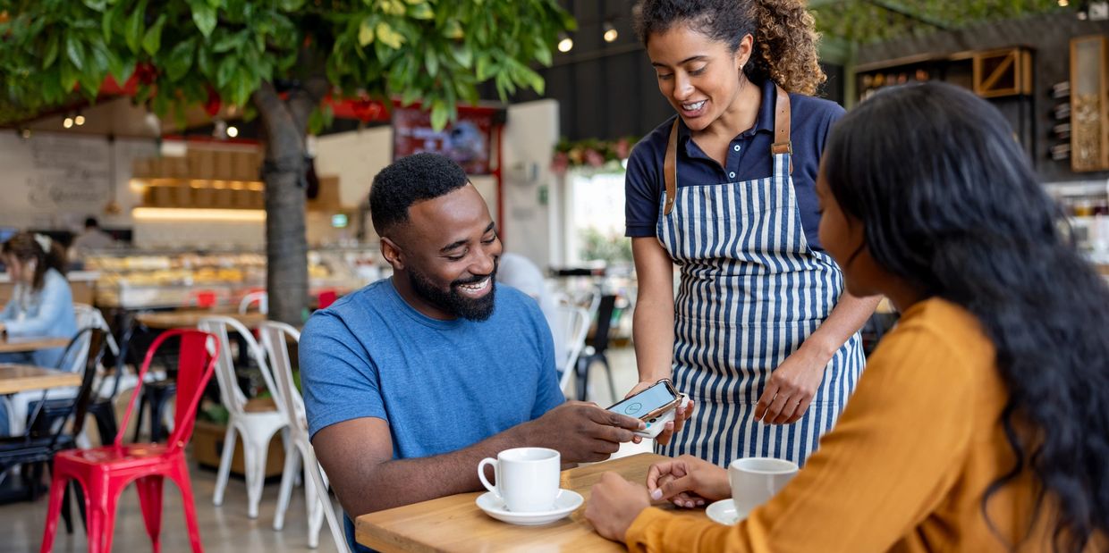 A waiter helping customers pay by phone at a cafe.