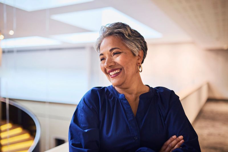 Close-up of a smiling mature woman with short grey hair and a blue blouse, arms crossed, in a bright, modern interior. Focus on her joyful expression.