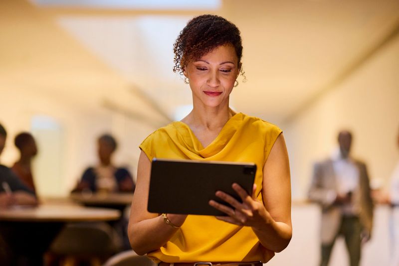 Focused woman in a yellow top reviews content on her tablet, bathed in warm light. Blurred figures suggest a collaborative office environment.