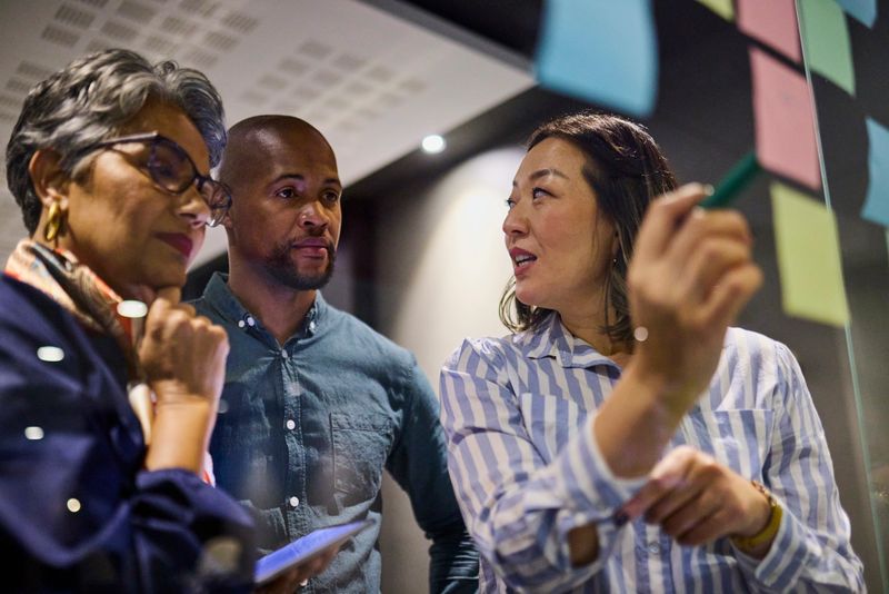 Diverse group of three professionals collaborating on ideas written on sticky notes on a glass wall. Focus on the woman writing, with others attentively listening. Bright, modern office setting.