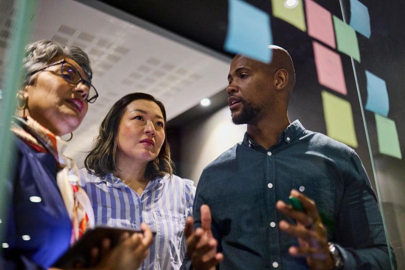 Low-angle shot of three diverse professionals engaged in a brainstorming session, reviewing colorful sticky notes on a glass board. The man gestures while the women listen attentively.