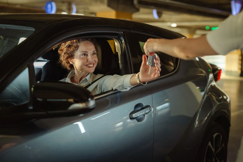 A happy individual receives car keys from another person while seated in a compact car, showcasing automotive transactions or rental services in a covered parking environment.