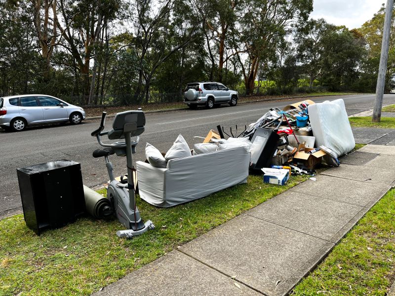 A large pile of rubbish set out on the curb for council collection in a suburban neighbourhood. The heap includes oversized items like flattened cardboard boxes, a worn couch with pillows, several mattresses stacked atop one another