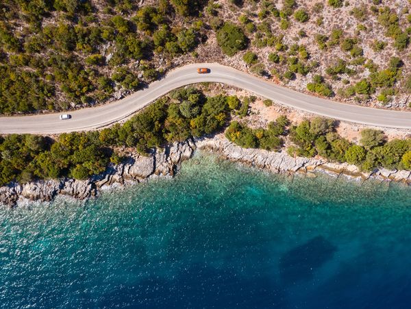 A winding coastal road with cars beside clear blue water and rocky shore.