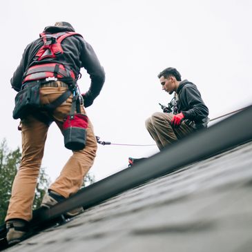 Two workers on a roof wearing safety gear and harnesses.