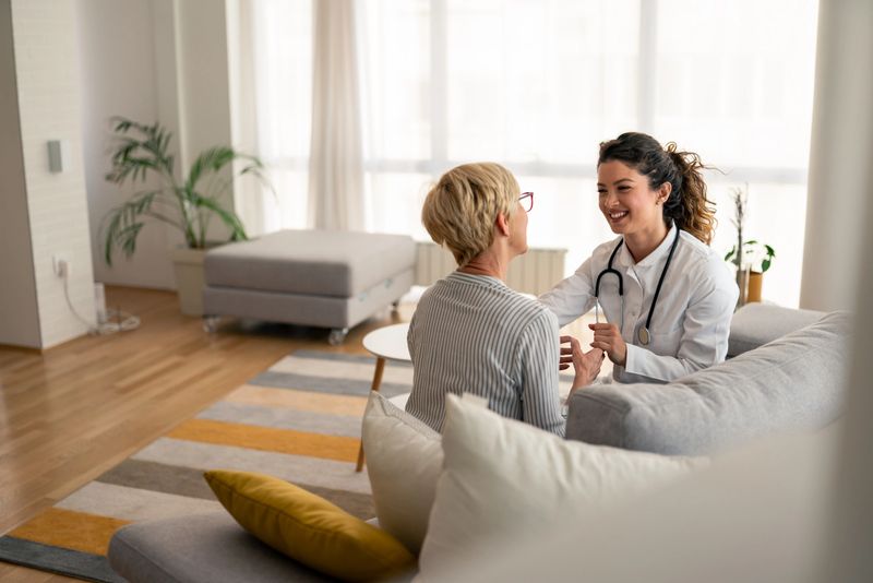 Caucasian female physician conducts friendly medical consultation with senior patient, wearing white coat and stethoscope during residential healthcare visit.