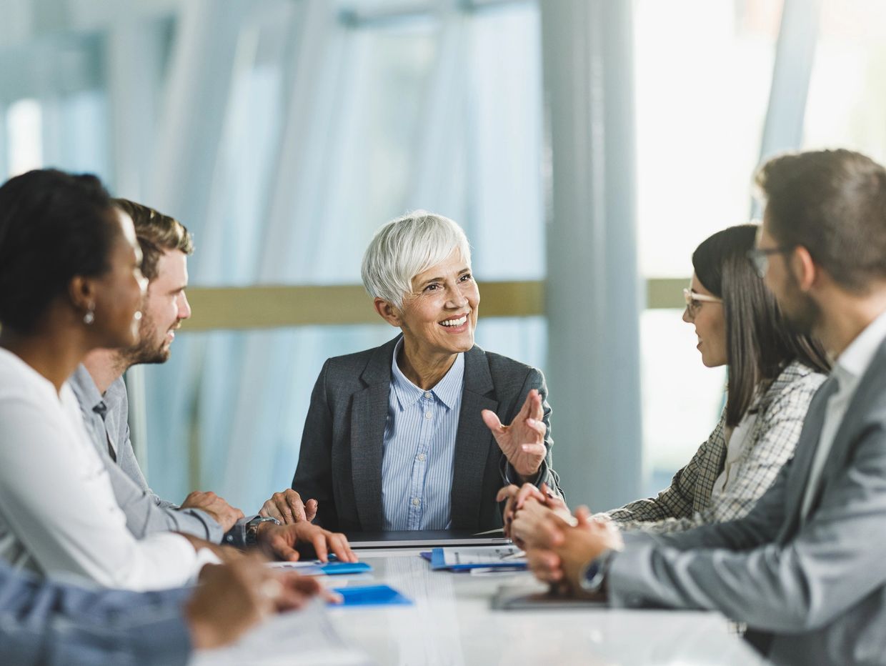 A senior woman leads a business meeting with engaged colleagues around a table.