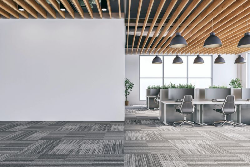 A modern office interior with an open floor plan. On the left, there is a blank white wall with copy space, and on the right, rows of workstations with computers, chairs, and greenery are illuminated by large windows and pendant lights. The ceiling features wooden slats, complementing the gray patterned carpet flooring. 3D rendered image.