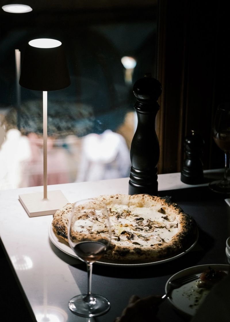 An inviting scene in an Italian restaurant featuring a freshly baked 4 Formaggi pizza on a wooden plate, paired with a glass of wine and a pepper mill on a window-side table. The setting is softly lit by a sleek table lamp, creating a warm and cozy dining atmosphere. The perfect blend of melted cheeses on the pizza highlights the rich and authentic flavors of Italian cuisine, making this an ideal setup for an intimate or gourmet dining experience.