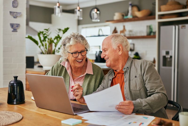 Smiling senior couple discussing bills and reviewing documents while using laptop at home. Mature couple collaborating on financial management at their dining table. Elder man and retired woman sharing a moment while organizing their finances while using computer.