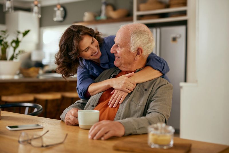 Smiling senior father and daughter embracing while enjoying tea at home. Elderly father and adult daughter bonding over tea and conversation. Warm moment between father and daughter sharing tea mugs while looking at each other.
