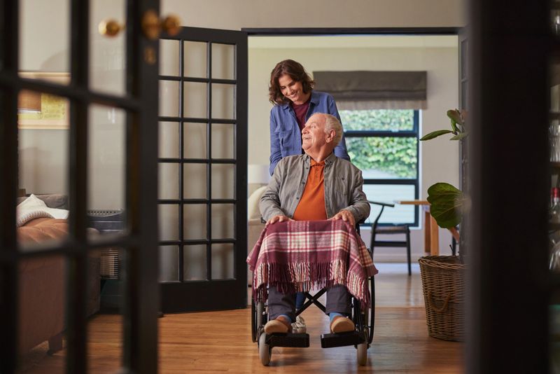 Senior man sitting on a wheelchair while his daughter assists him for a ride. Lovevly daughter supporting her old father by helping with his wheelchair indoors. Happy smiling caregiver woman visiting and supporting disabled elderly man at home.
