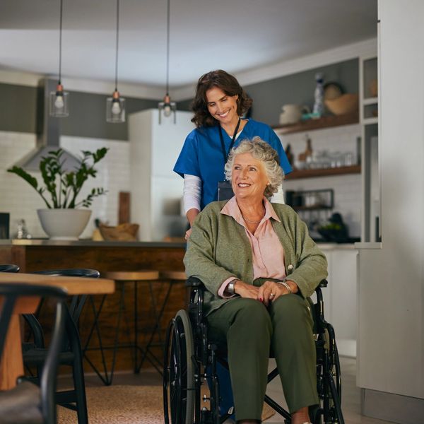 Caregiver smiling and assisting a happy senior woman in a wheelchair inside a home.