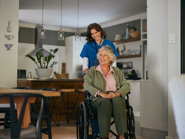 A caregiver in blue scrubs pushes a smiling elderly woman in a wheelchair through a cozy kitchen.