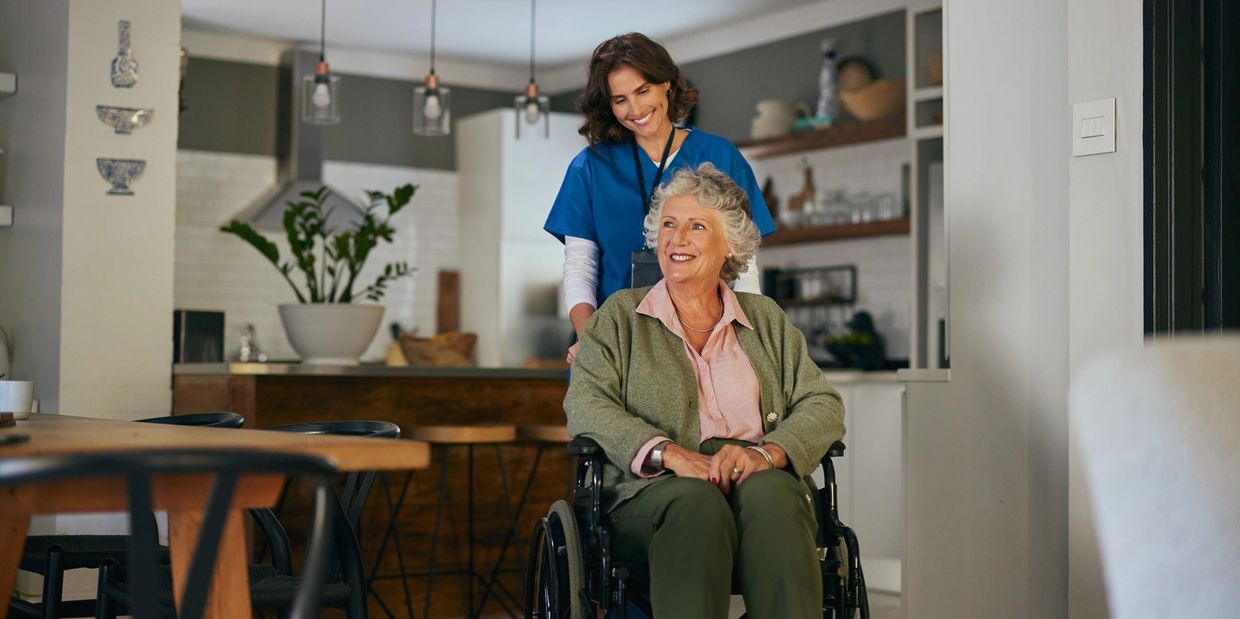 Caregiver smiling while pushing elderly woman in wheelchair at home.