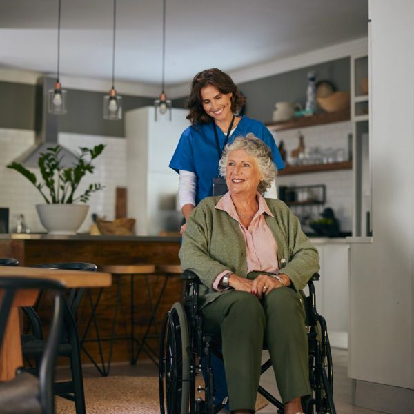 Caregiver pushing elderly woman in wheelchair, both smiling in a cozy home kitchen.