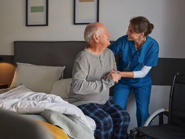 Nurse assisting elderly man sitting on a bed, smiling warmly.