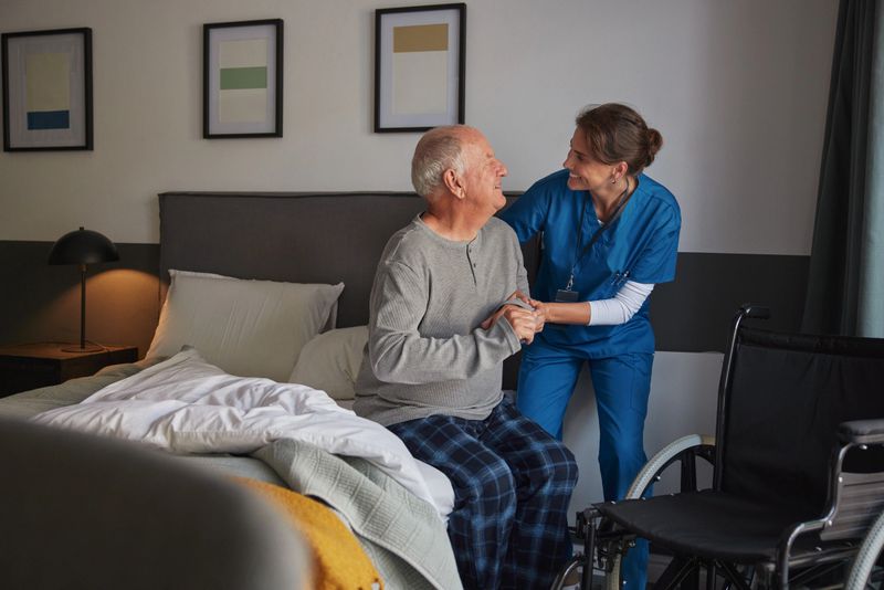 Caring nurse helping an elderly man in pajamas get up from bed in a bedroom. Mid adult healthcare professional supporting senior man while getting up from bed and move towards wheelchair at home. Compassionate caregiver providing support to old man during morning routine.