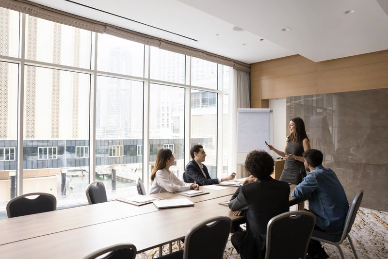 Skilled European female leader coaching interested business team, giving educational workshop, making flip chart presentation in boardroom. Staff members listening to professional coach at workshop