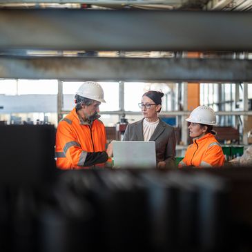 Two workers in orange safety gear and a businesswoman discuss over a laptop in an industrial setting.