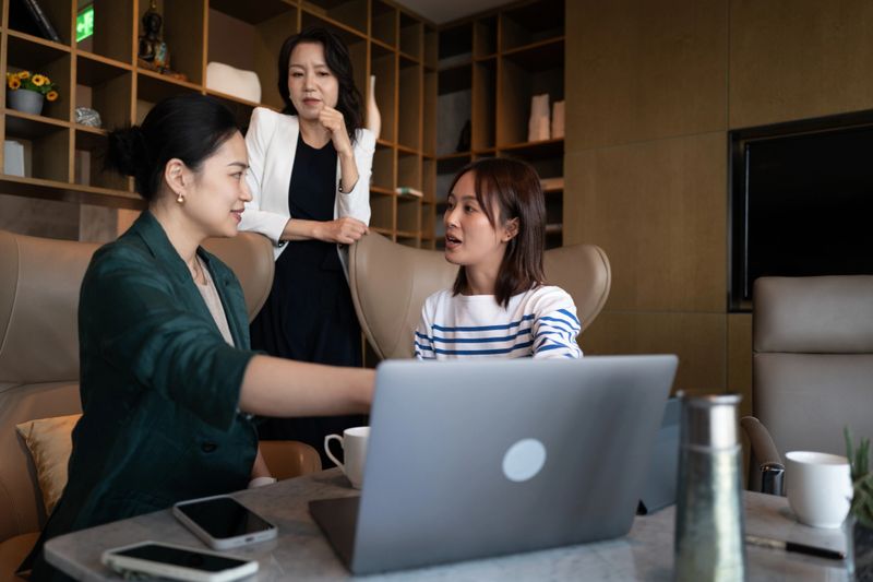 A female programmer is presenting new application features on her laptop to two female managers in a modern office lounge. The scene captures the interaction, discussion, and collaboration involved in showcasing a new product or technology. The managers are listening attentively and engaging with the demonstration. Shot with a Leica Q3 camera.
