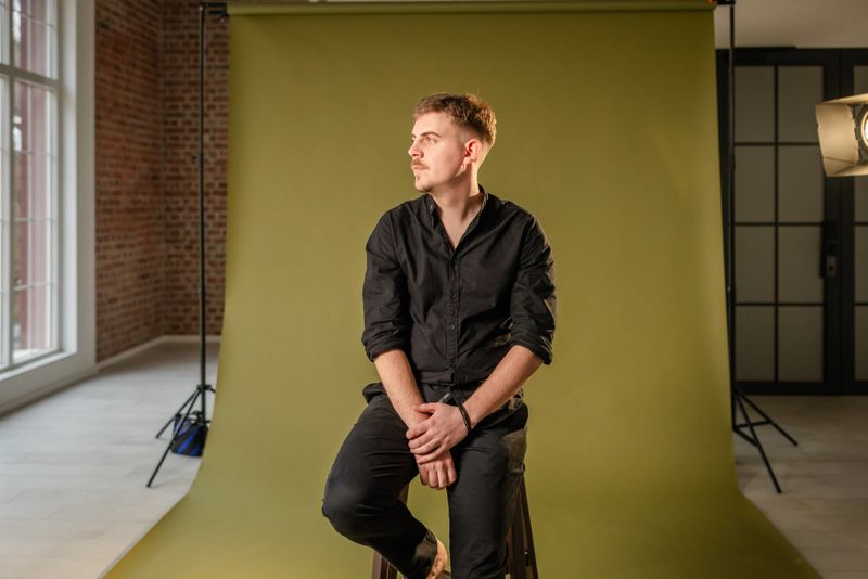 A casually dressed man enjoys his time in a photo studio, posing on a stool with a professional lighting setup and a green backdrop, conveying artistic creativity.