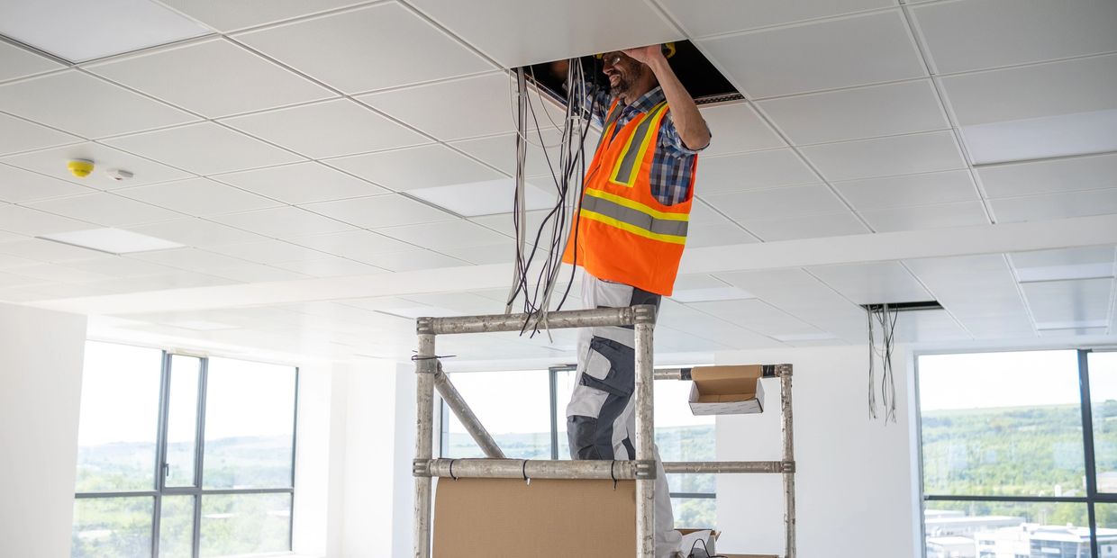 Electrician installing wiring in office ceiling while standing on scaffolding.