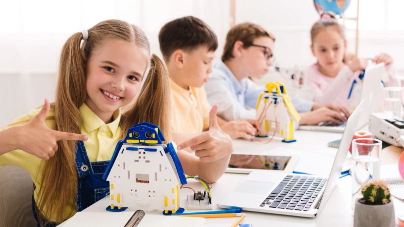 Students participate in a robotics workshop at a well lit classroom, focused on building their projects. A girl smiles proudly with her robot while peers are concentrated on their own creations.