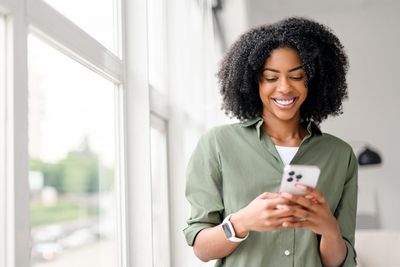 Smiling woman looking at her smartphone near a bright window.