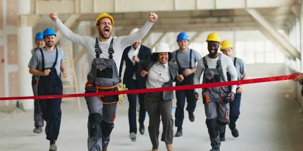 Construction workers and a businesswoman racing to break a finish line ribbon indoors.