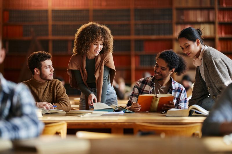 Multiracial group of happy university students cooperating while learning a lecture from books in library.