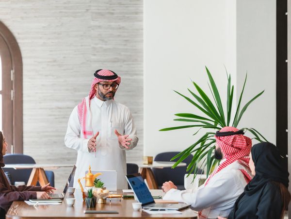 A man in traditional Middle Eastern attire speaking to colleagues in a modern office.
