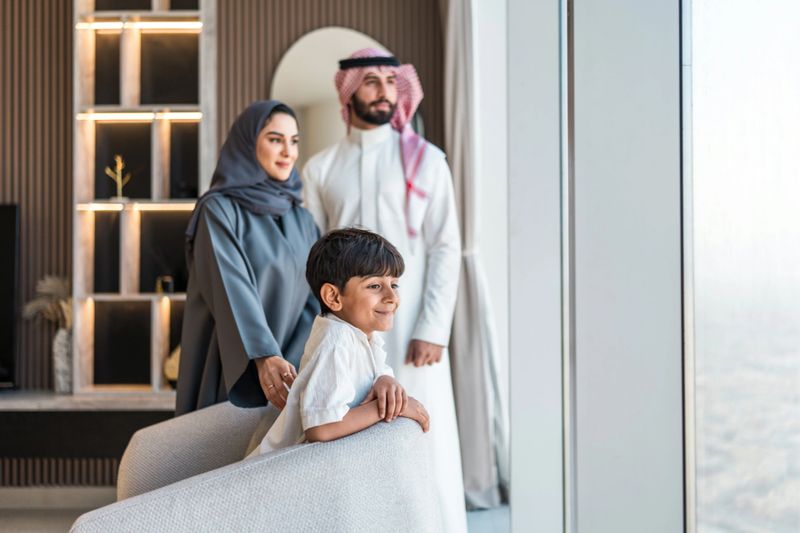 A diverse, joyful Arabic family admires the stunning cityscape of Riyadh through large windows in their contemporary home. The family, dressed in traditional attire, revels in the breathtaking view, portraying a serene and happy moment.