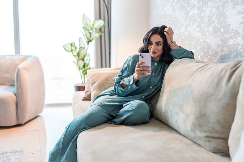 A young Arabic woman in elegant attire relaxes on a beige sofa, engaging with her smartphone. The modern home setting features soft natural lighting and minimalistic decor, including a potted plant by the window.