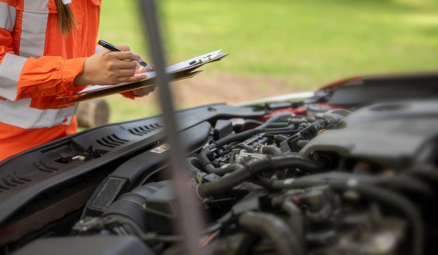 Person in orange jacket inspecting a car engine and taking notes on a clipboard.