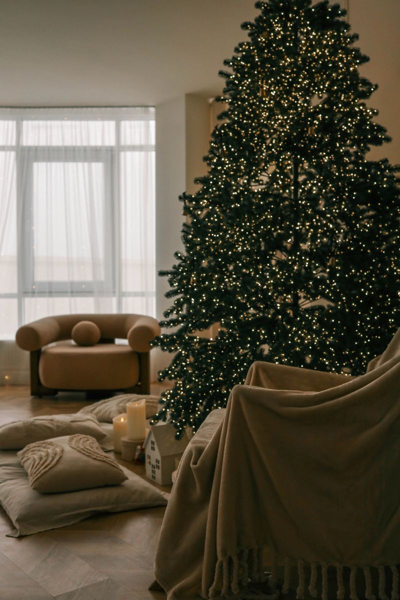 Cozy living room with a warm beige armchair, soft pillows, and a beautifully decorated Christmas tree glowing with fairy lights. The serene atmosphere is perfect for holiday relaxation.