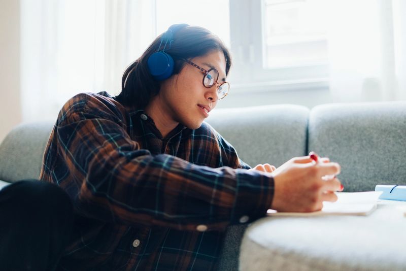 A young individual wearing headphones and glasses concentrates on writing notes while seated on a cozy couch at home.