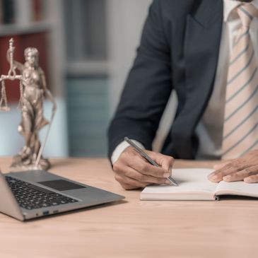 Lawyer writing notes at desk with laptop and justice statue.
