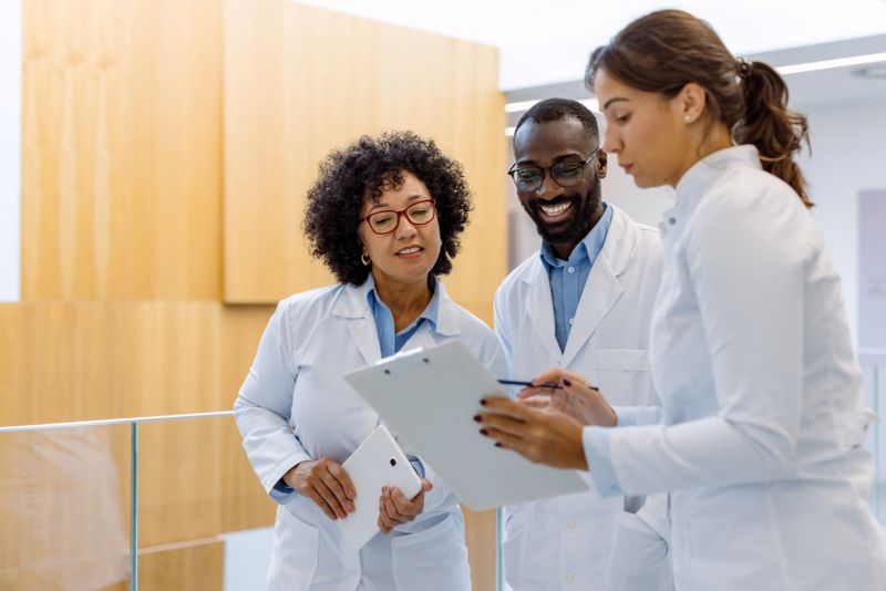 Three doctors are standing in a hospital hallway, reviewing patient information on a clipboard and discussing treatment options with a tablet in hand