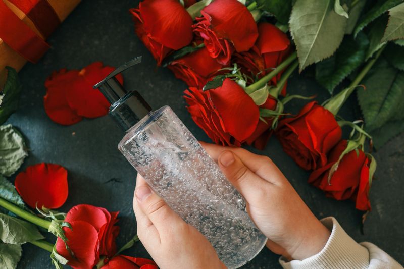 Transparent pump bottle with clear gel placed among vibrant red roses and petals on a dark textured surface, emphasizing a modern and fresh aesthetic.