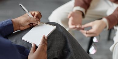 A person taking notes while another talks, holding glasses in hand.