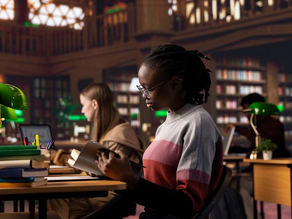 Students studying quietly in a library with green reading lamps and books.